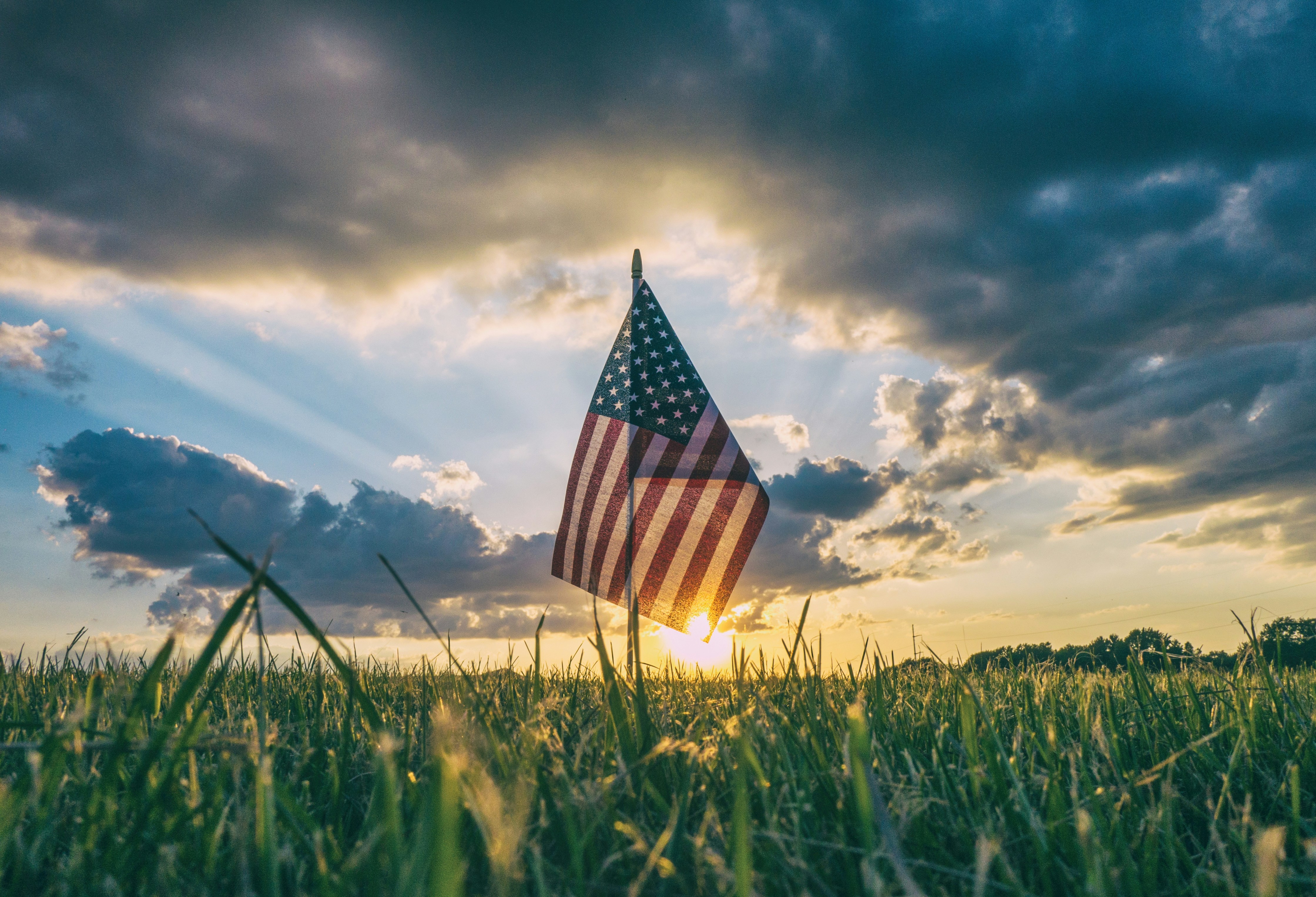 A solo U.S. flag staked in grass in the foreground touches a coming-sunset and clouds in the background