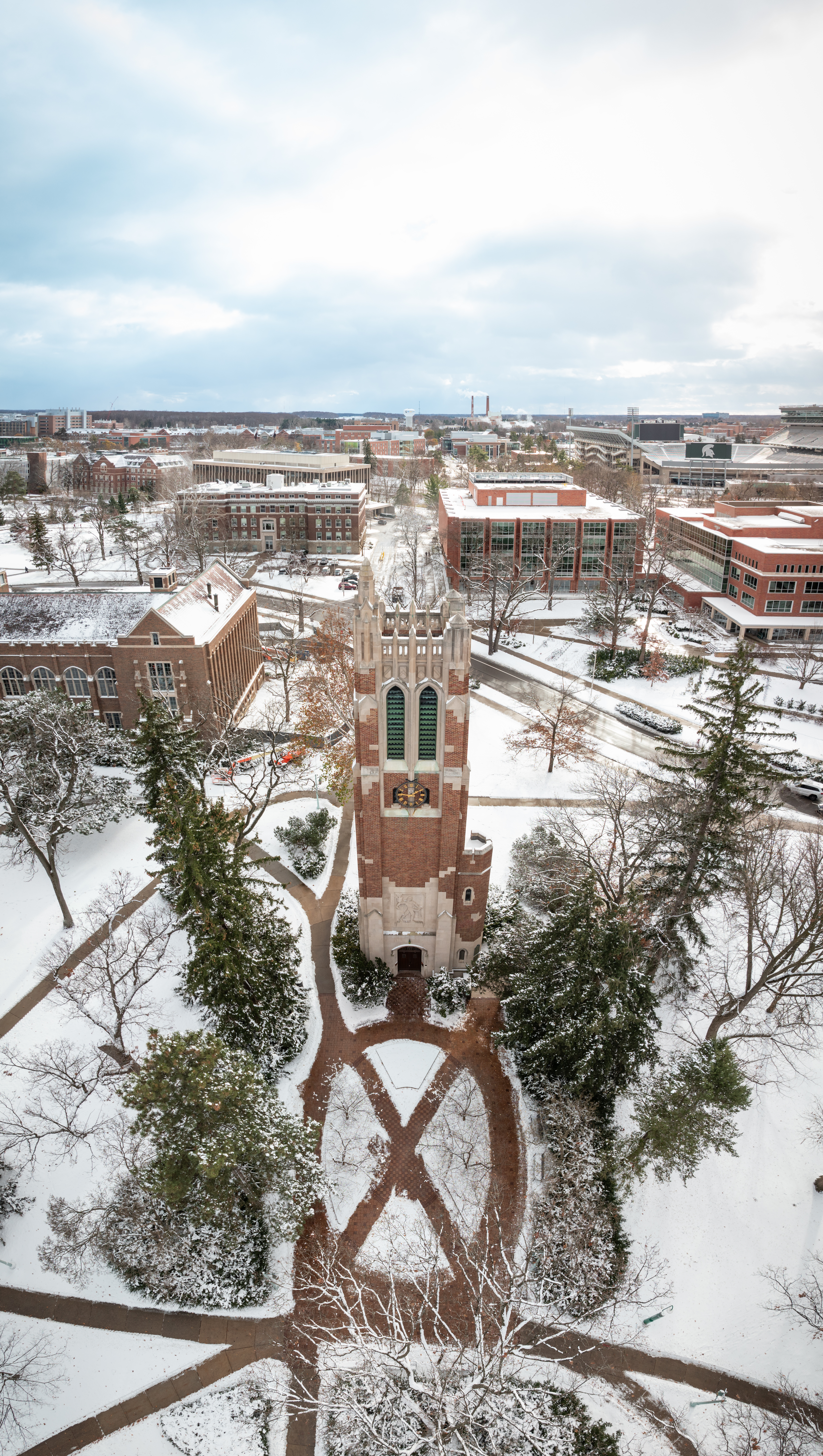 Aerial view of Beaumont Tower at the center of Michigan State University's campus after a snowfall. Snow-covered walkways, trees, and historic academic buildings surround the tower, with Spartan Stadium and additional campus buildings visible in the distance beneath a cloudy winter sky.