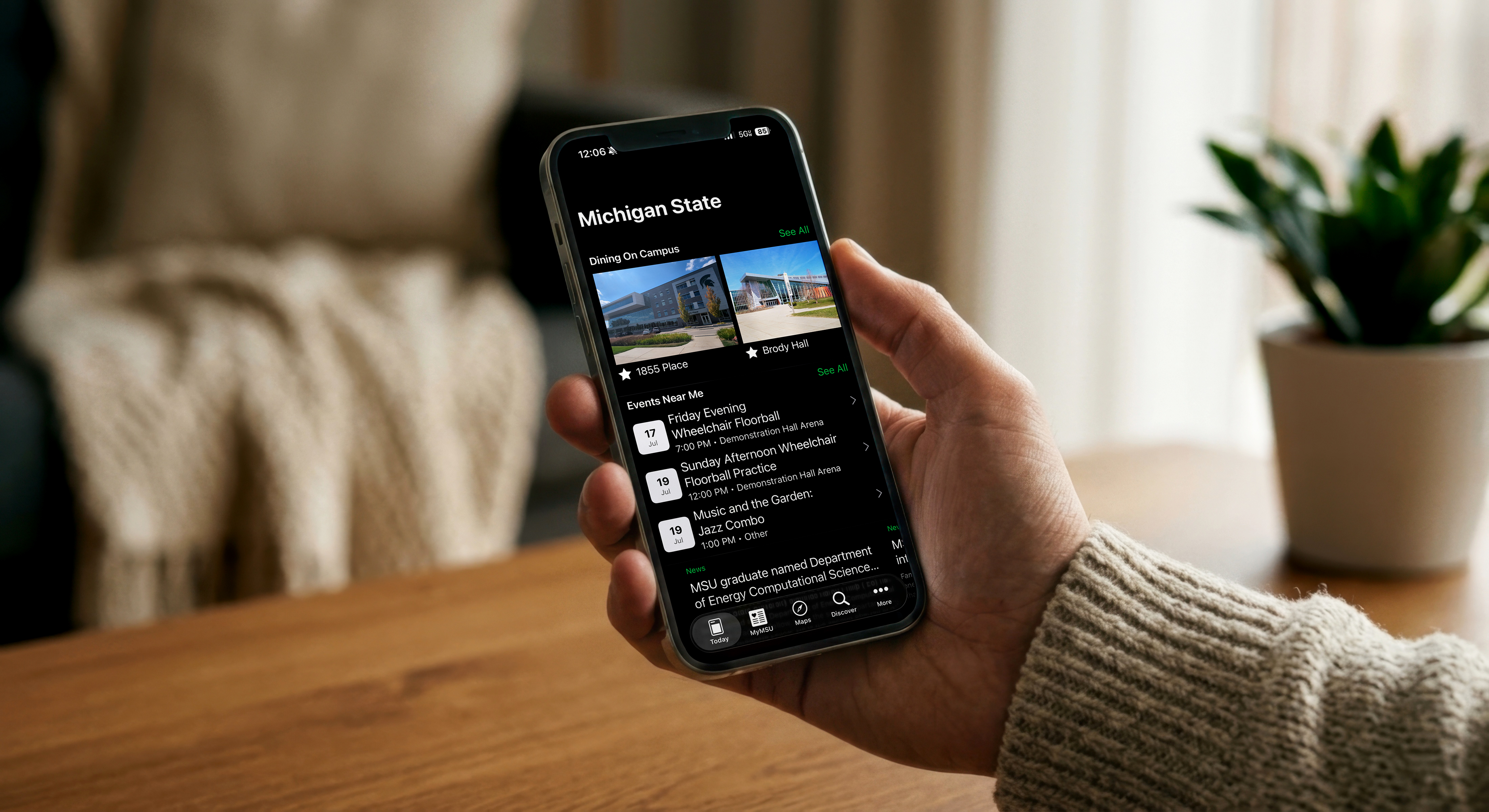 A person wearing a cream-colored knit sweater holds a smartphone displaying the Michigan State mobile app in dark mode. The app features campus dining locations, nearby events, and news, while the phone is in sharp focus against a softly blurred indoor background with a wooden table, bed, and potted plant.