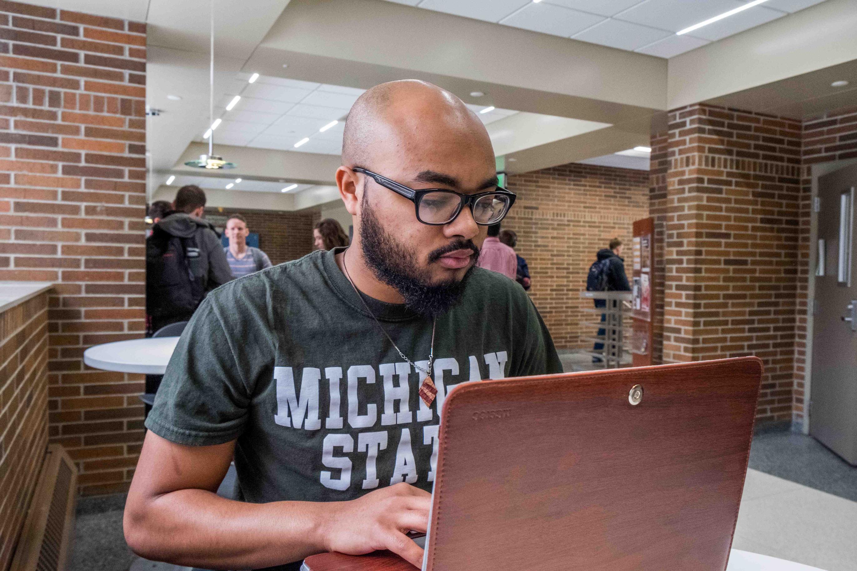 A Michigan State University student wearing a green MSU T-shirt works on a laptop while seated at a table inside a campus academic building. Other students gather and walk through the bright, open common area with brick walls and study spaces in the background.