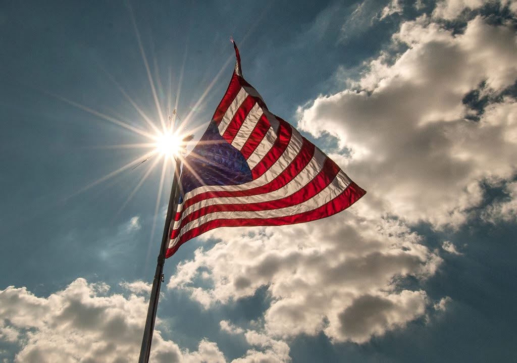 U.S. flag flies high on a pole under sun and clouds