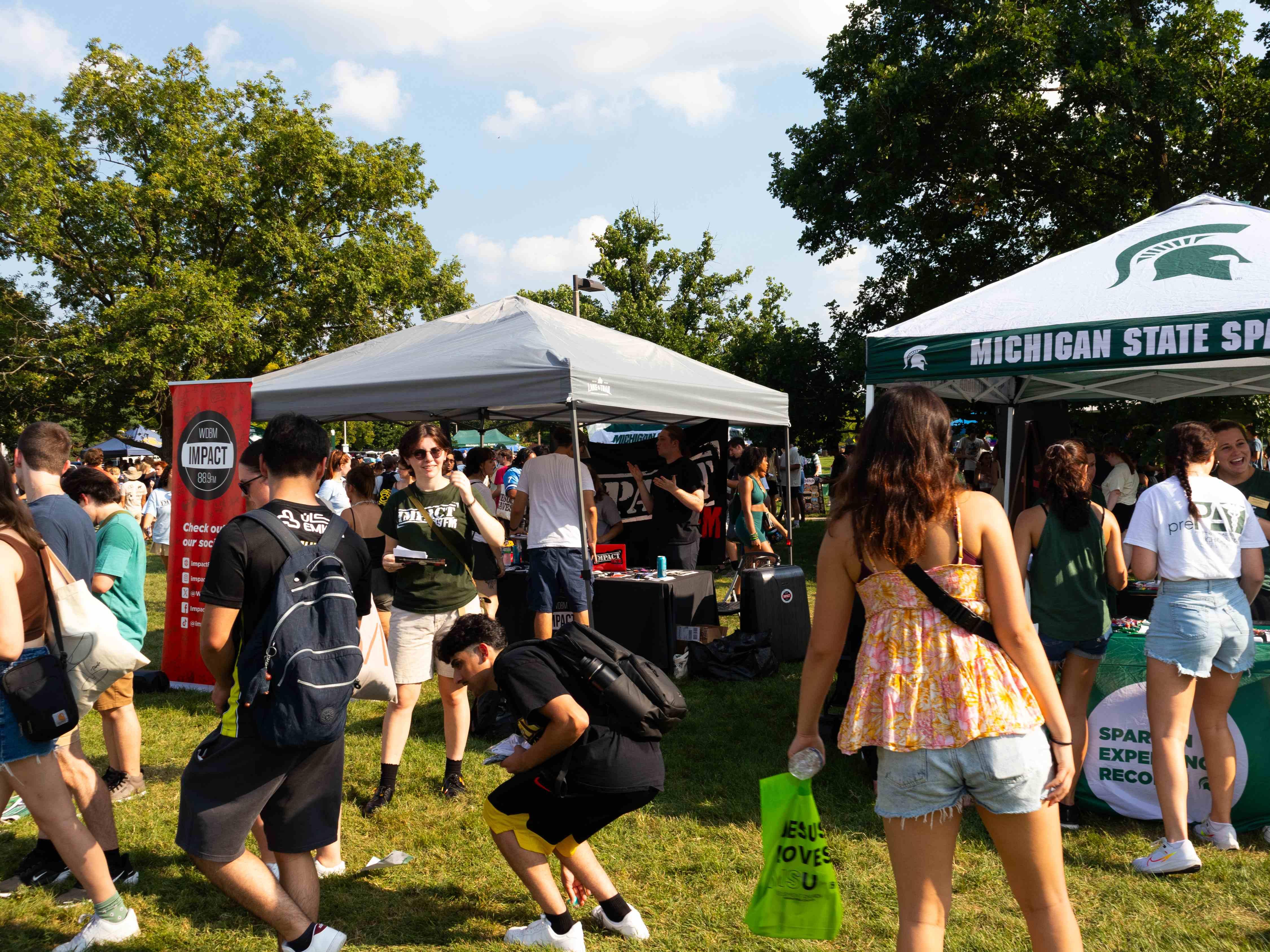 Students gather around Michigan State University tents during an outdoor campus involvement event on a sunny day. Staff members speak with students at information tables while attendees walk between booths, creating a busy and welcoming atmosphere on a grassy lawn surrounded by mature trees.