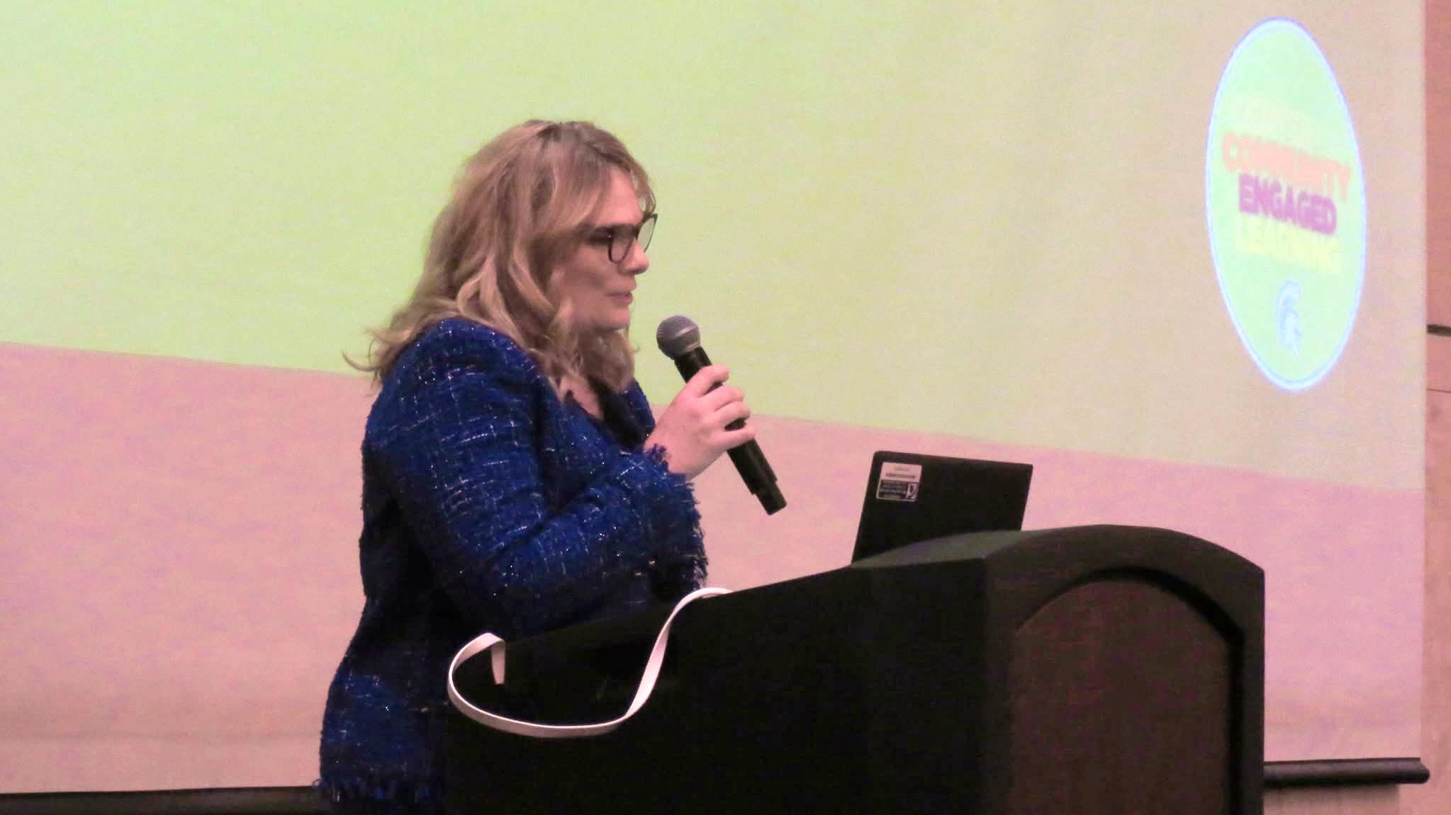 Renee Speaking at podium with screen presentation behind stage setup