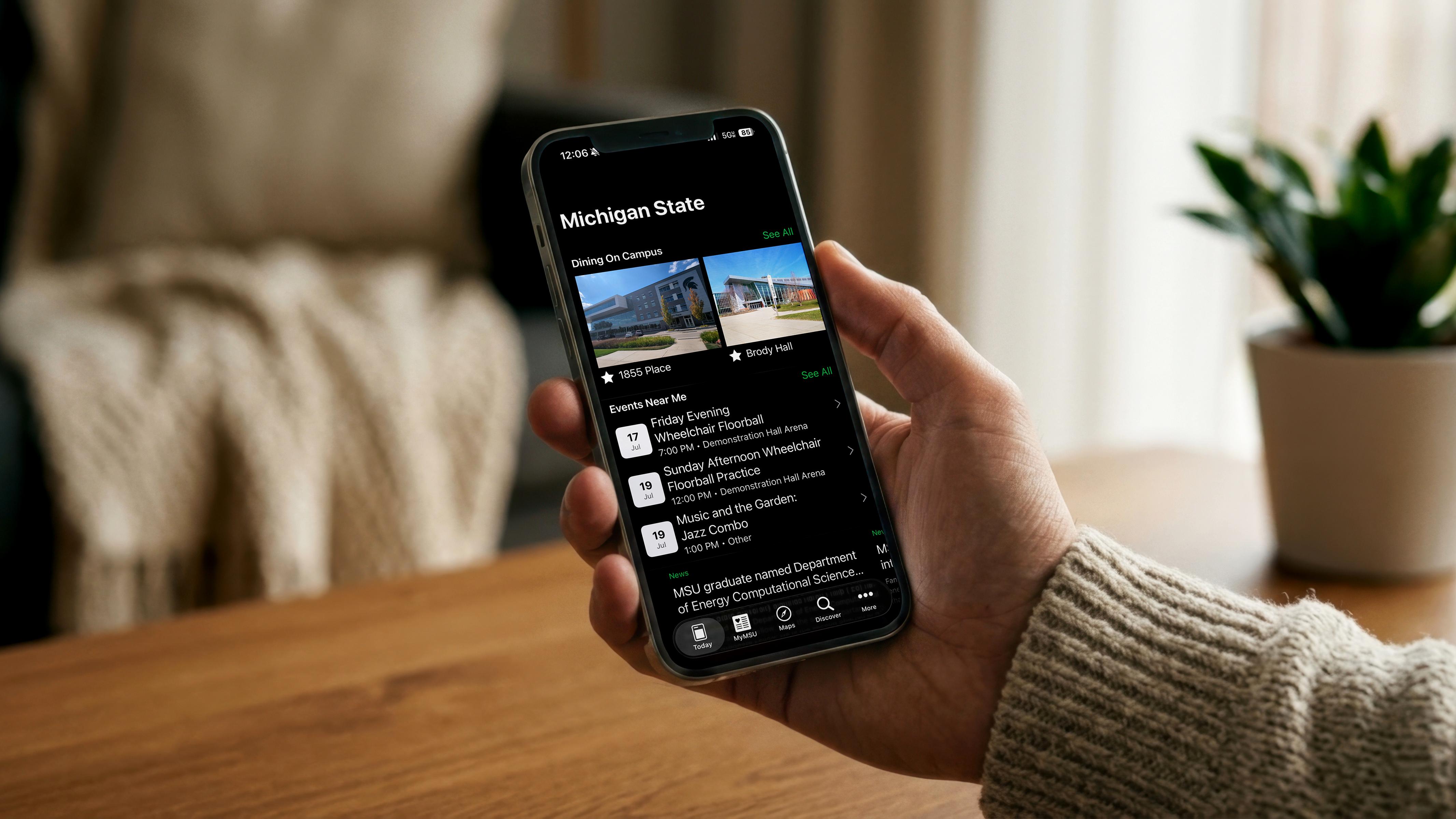 A person wearing a cream-colored knit sweater holds a smartphone displaying the Michigan State mobile app in dark mode. The app features campus dining locations, nearby events, and news, while the phone is in sharp focus against a softly blurred indoor background with a wooden table, bed, and potted plant.