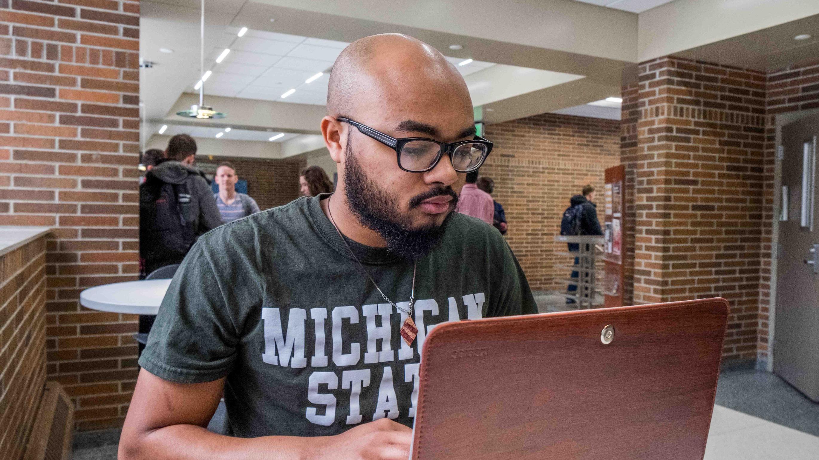 A Michigan State University student wearing a green MSU T-shirt works on a laptop while seated at a table inside a campus academic building. Other students gather and walk through the bright, open common area with brick walls and study spaces in the background.