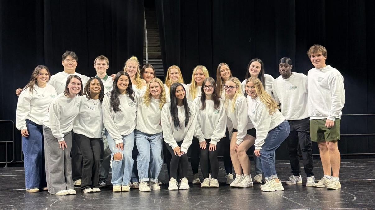 A group of students wearing white shirts and jeans stand on a stage