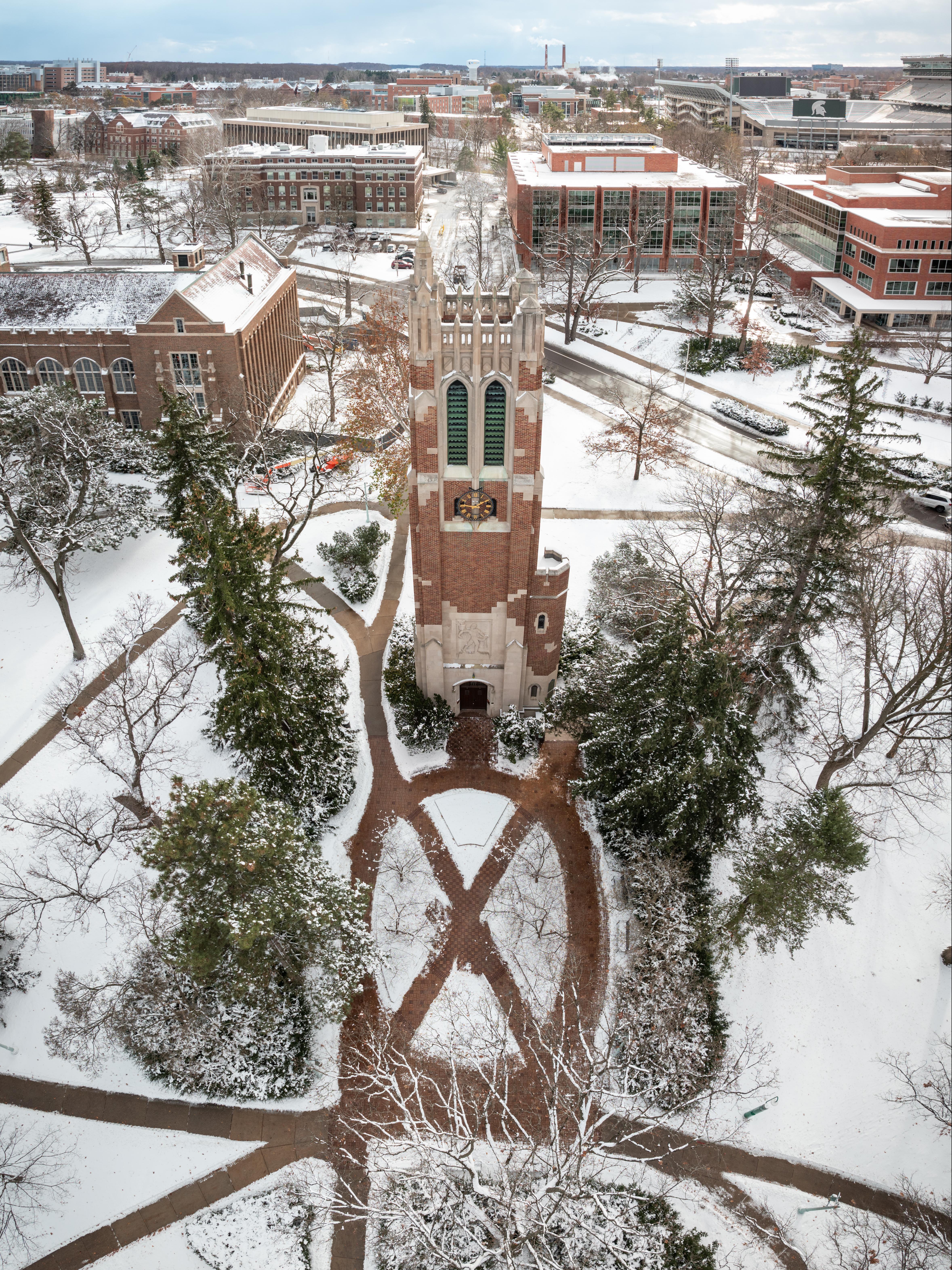 Aerial view of Beaumont Tower at the center of Michigan State University's campus after a snowfall. Snow-covered walkways, trees, and historic academic buildings surround the tower, with Spartan Stadium and additional campus buildings visible in the distance beneath a cloudy winter sky.